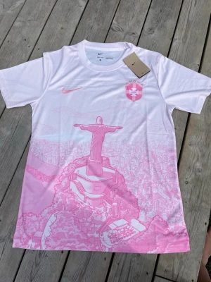 Vit och rosa Nike Brasilien fotbollströja  - Säljer en snygg vit fotbollströja från Nike med rosa detaljer och motiv av Kristusstatyn i Rio de Janeiro. Tröjan har korta ärmar, rund hals och klubbmärke på bröstet. Perfekt för dig som gillar unika sportkläder och cool design.