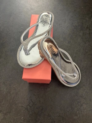 Silvriga sandaler med strassdetaljer - Snygga sandaler från SIMMI i spegelblankt silver med tunna remmar dekorerade med strass. Platt sula och öppen tå ger en trendig look. Perfekta för dig som vill sticka ut med lite extra bling till sommaren. Skorna är dessutom helt nya och oanvända.