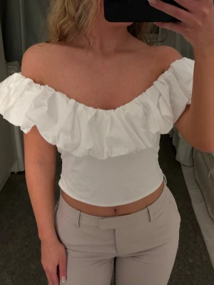 Vit offshoulder topp med volang - Säljer en snygg vit offshoulder topp med stor volang runt hela halsen. Toppen är croppad och har en tight passform som framhäver figuren. Perfekt för dig som vill ha en trendig och stilren look.