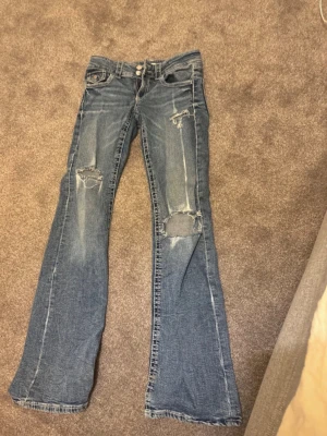 Blå slitna bootcut jeans - Ett par blå bootcut jeans med slitningar och hål på låren och knäna. Jeansen har låg midja och klassisk femficksdesign. Perfekta för en avslappnad och trendig look. Strl 158