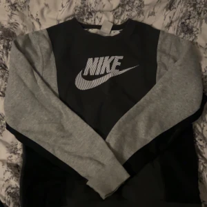 Nike sweatshirt med grå ärmar - Säljer en snygg svart sweatshirt från Nike med kontrasterande ljusgrå ärmar och stort vitt Nike-logotryck på bröstet. Perfekt för dig som gillar sportiga och stilrena plagg. Tröjan har en klassisk rund hals och långa ärmar.