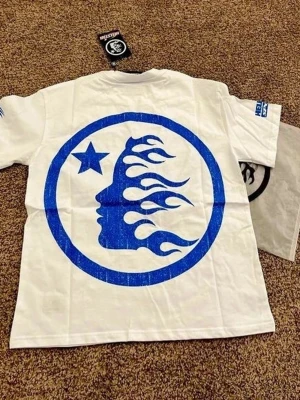 Vit t-shirt med blått tryck från HELLSTAR - Vit t-shirt från HELLSTAR med stort blått tryck på ryggen som föreställer en profil med eldflammor och en stjärna. Klassisk passform och rund hals. Tryck även på ena ärmen. Perfekt för dig som gillar streetwear och grafiska motiv.