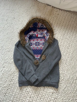 Grå hoodie med fejkpäls - Mysig grå hoodie med fluffig fuskpäls runt luvan och färgglatt vintermönster i blått, vitt och rött på luvans insida. Dragsko med tofsar och långa ärmar. 