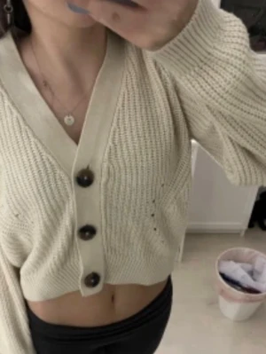 Beige cropped kofta med stora knappar - Trendig beige stickad kofta med stora svarta knappar framtill och bred ribbad kant. Croppad modell med lång ärm och v-ringad hals. Perfekt att styla med höga jeans eller kjol.