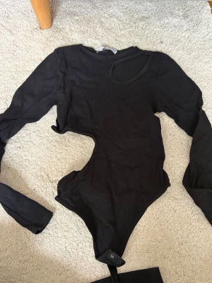 Svart ribbad body med cut-out - Trendig svart body med lång ärm och cool cut-out detalj vid axel och midja. Tillverkad i ribbat stretchigt material som sitter snyggt på kroppen. 