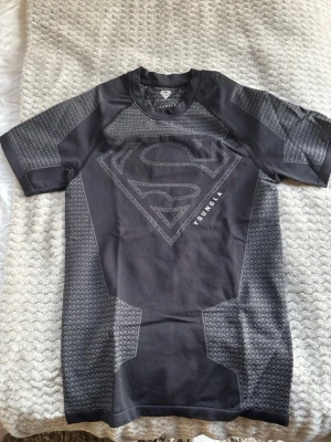 Svart kompressionströja Superman YoungLA - Snygg svart kompressionströja från YoungLA med Superman-logga på bröstet. Tröjan har korta ärmar, tight passform och mönstrade paneler i grått. Perfekt för gymmet eller träning, tillverkad i stretchigt funktionsmaterial.