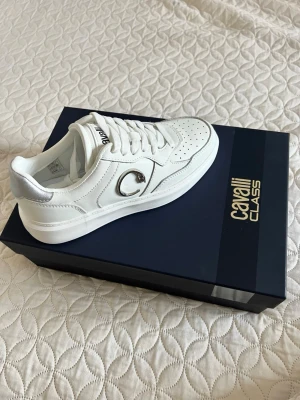 Vita Cavalli Class sneakers storlek 37  - Stilrena vita sneakers från Cavalli Class av Roberto Cavalli med silverdetaljer, snörning samt diskret logga på plösen och hälen. Väldigt lätta skor att ha på sig då de knappt väger något. Dom är aldrig använda då de aldrig kommit till användning. Nypris: ca 4500kr och kommer i originalkartongen. 