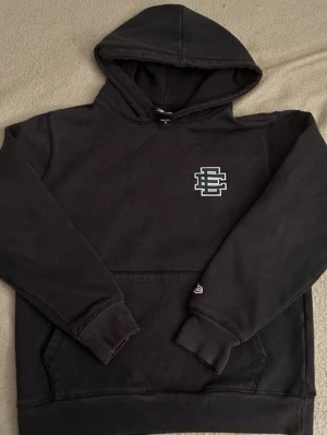 Svart hoodie med vit logga - Svart hoodie från New Era med vit broderad logga på bröstet. Klassisk känguruficka framtill och ribbade muddar vid ärmslut och nederkant. Perfekt för en avslappnad streetwear-look. Materialet är mjukt och bekvämt bomullstyg.