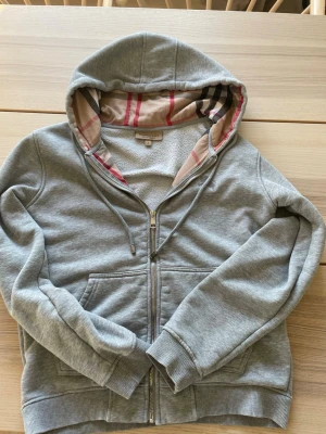 Grå zip-hoodie från Burberry - Säljer en grå hoodie från Burberry med dragkedja och klassiskt rutigt foder i huvan. Hoodien har broderad logga på bröstet, två fickor fram och ribbade muddar. Perfekt för dig som gillar stilrena och exklusiva plagg med snygga detaljer.