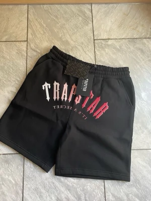 Svarta shorts från Trapstar - Svarta shorts från Trapstar med tryckt logga i rött och vitt framtill. Modellen har elastisk midja och sidofickor. Perfekta för en avslappnad streetwear-look.