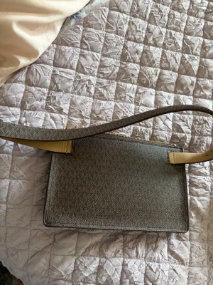 Brun midjeväska från Michael Kors - Snygg brun midjeväska från Michael Kors med klassiskt MK-mönster. Väskan har en rektangulär form,  och en elegant guldfärgad kedja som detalj. Perfekt för dig som gillar stilrena accessoarer med lyxig känsla.Äkta