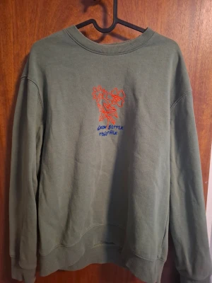 Grön sweatshirt med blommor - Cool grön sweatshirt med tryck av orange blommor och texten 'Grow Better Together' i blått på bröstet. Tröjan har rund hals, långa ärmar och ribbade muddar. Perfekt för dig som gillar en avslappnad och trendig stil.