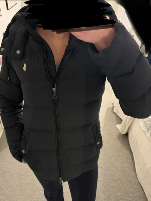 Svart dunjacka från Polo Ralph Lauren - Säljer en svart dunjacka från Polo Ralph Lauren med klassisk logga på bröstet. Jackan har huva, dragkedja framtill och två fickor med knappstängning. Den är i fint skick men har ett litet hål som inte är synligt❤️(bild 3)