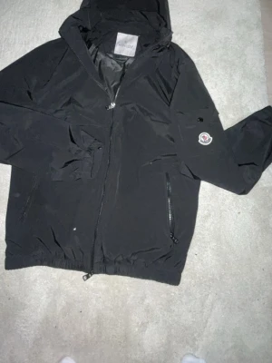 Moncler windbreaker - Snygg svart vindjacka från Moncler med huva och dragkedja framtill. Jackan har två fickor med dragkedja och Moncler-logga på ärmen. Insidan har ett unikt serietidningsmönster. Tillverkad i lätt och slitstarkt material, perfekt för blåsiga dagar. Pris kan diskuteras vid snabb affär.