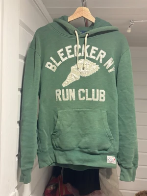 Grön hoodie Polo Ralph Lauren M - Grön hoodie från Polo Ralph Lauren med trycket 'BLEECKER NY RUN CLUB' och vingar framtill. Klassisk känguruficka, huva med dragsko och ribbade muddar. Perfekt för en avslappnad streetstyle-look.
