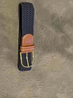 Mörkblått flätat bälte med läderdetalj - Snyggt mörkblått flätat bälte med bruna läderdetaljer och guldfärgat spänne. Perfekt accessoar för att lyfta din outfit med en klassisk touch. Bältet har en flexibel design som gör det enkelt att justera efter behov.obs går att upphämtas i södra Stockholm 
