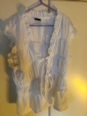 Vit blus med volanger och knyt - Superfin vit blus från Gina Tricot med volangdetaljer och knyt framtill. Blusen är ärmlös och har en luftig, lätt struktur i bomull. 