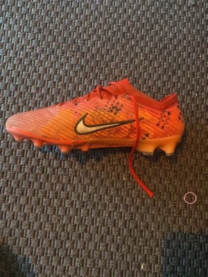 Nike fotbollsskor  - Säljer ett par snygga orangea Nike fotbollsskor med svart och vit swoosh på sidan. Skorna har snörning, låg profil och mönstrad ovandel i syntetmaterial. Perfekta för spel på gräsplan. Detaljer i svart och små stjärnmönster på ovandelen.