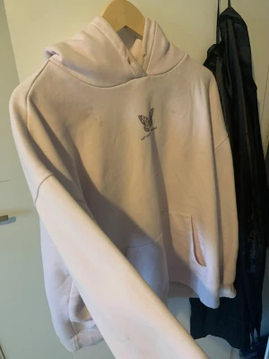 Ljusrosa hoodie med tryck - Ljusrosa hoodie med huva och känguruficka. På bröstet finns ett litet fjärilstryck och texten 'LET YOUR SOUL'. Baksidan har ett stort grafiskt tryck med sol, måne och texten 'FREE YOUR SOUL'. Perfekt för dig som gillar unika detaljer och streetwear-vibe.