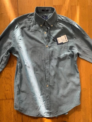 Blå linneskjorta från Gant - Helt ny oanvänd Snygg blå skjorta från Gant i linne med klassisk button-down krage och bröstficka. Skjortan har långa ärmar och vita knappar framtill. Perfekt för en stilren och avslappnad look.