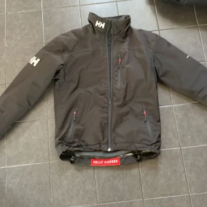 Svart vindjacka Helly Hansen - Svart vindjacka från Helly Hansen med HH-logga på ärm, bröst och krage. Jackan har dragkedja framtill, tre fickor med dragkedja och detaljer i rött. Insidan är fodrad med mjukt material och kragen är hög för extra skydd. Perfekt för blåsiga dagar. Passar perfekt för dem som är 173-185 långa 