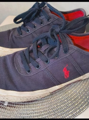 Mörkblå Polo Ralph Lauren sneakers - Snygga mörkblå sneakers från Polo Ralph Lauren med klassisk röd logga på sidan. Skorna har lågt skaft, blå snören och röd-orange innersula. Ovandelen är i canvas och sulan är vit. Perfekta för en stilren och avslappnad look. Se bilder för bedömnig. Storlek 43 pris kan diskuteras