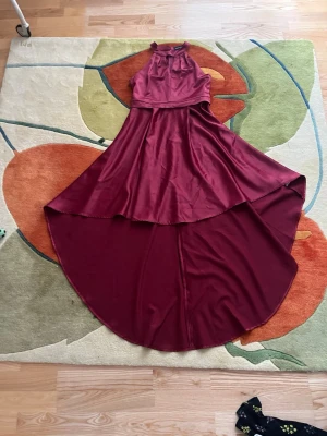 Vinröd långklänning i satin - Snygg vinröd långklänning i glansigt satinliknande material med hög hals och ärmlös design. Klänningen har en asymmetrisk fåll med längre bakstycke och en elegant, flytande siluett. Perfekt för dig som vill sticka ut med en stilren look.