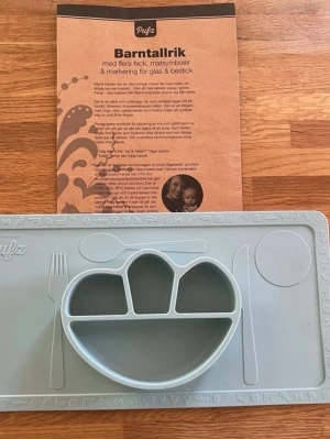 Pufz Barntallrik med flera fack - Barntallrik med underlägg är tillverkad av livsmedelsgodkänd silikon och är dekorerad med symboler och markeringar för glas och bestick. Barntallriken med egen bricka passar Ikeas klassiska barnstol Antilop som hand i handske, men fungerar lika bra direkt på matbordet.  Färg: turkos/ grå  Tål maskindisk Kan användas i frys, ugn och mikrovågsugn Klarar temperaturer från -50 °C till 230 °C 100 % giftfri livsmedelsklassad silikon (BPA free) Storlek: 21 x 38 cm