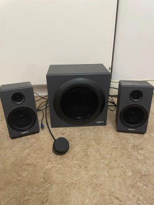 Logitech trådlösa högtalare svart - Säljer ett par stilrena trådlösa högtalare från Logitech i svart färg. Setet består av en större subwoofer och en mindre högtalare, båda med mattsvart finish och rundade detaljer. Perfekt för att få kraftfullt ljud till datorn eller mobilen.