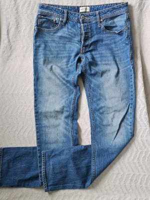 Blå slim fit jeans från Jack & Jones - Snygga blå slim fit jeans från Jack & Jones med klassisk femficksdesign och dragkedjegylf. Jeansen har en tvättad look med ljusare partier och kontrastsömmar. Perfekta för en avslappnad och trendig stil.