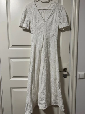 Vit långklänning med spetsdetaljer - Elegant vit långklänning med v-ringning och korta ärmar. Klänningen har vackra spetsdetaljer och ett diskret broderat mönster över hela tyget. Perfekt för sommaren eller för studenten 