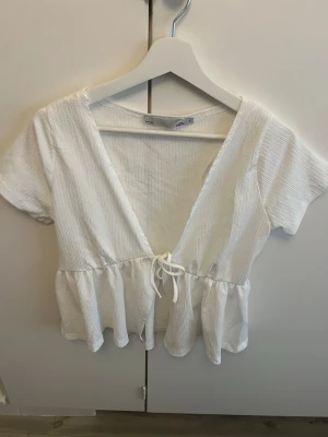 Vit ribbad blus från 157 - Söt vit ribbad blus från 157 med korta ärmar, v-ringning och knyt i midjan. Toppen har en lätt volang nedtill och är tillverkad i ett mjukt bomullsmaterial. Perfekt för en fräsch och enkel look.