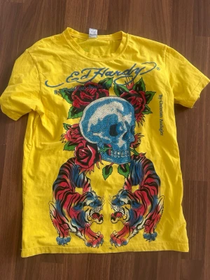 Gul Ed Hardy t-shirt med tryck - Fet gul t-shirt från Ed Hardy med stora, färgglada tryck av dödskalle, rosor, tigrar och drake. Klassisk crewneck och korta ärmar. Perfekt statement-plagg för dig som gillar unika motiv och streetstyle. Materialet är mjuk bomull.