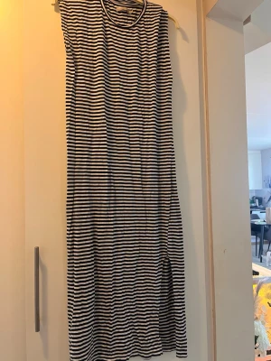 Randig långklänning i bomull - Säljer en svartvit långklänning med smala horisontella ränder. Klänningen är ärmlös och har en rak, avslappnad passform. Tillverkad i mjuk bomull, perfekt för varma dagar. Enkel och stilren design som passar till många tillfällen.