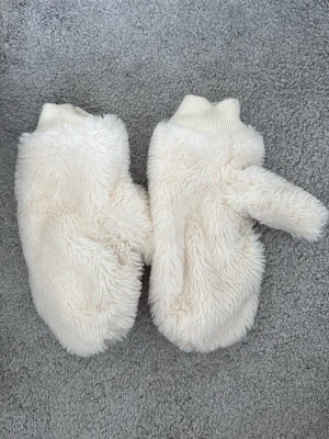 Vita fluffiga tumvantar i fuskpäls - Supermjuka och fluffiga vita tumvantar i fuskpäls med ribbad mudd. Perfekta för kalla dagar när du vill hålla händerna varma och samtidigt se stylish ut. Passar till de flesta vinteroutfits och är riktigt mysiga att ha på sig.