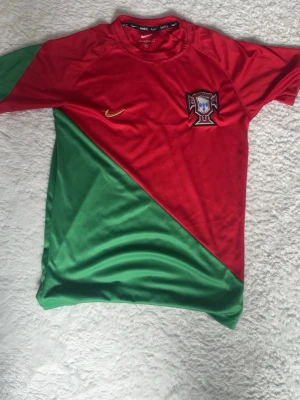 Portugal Nike fotbollströja röd/grön - Snygg Portugal fotbollströja från Nike i rött och grönt med diagonal färgdelning. Tröjan har broderad Nike-logga i gult och Portugals landslagsemblem på bröstet. Perfekt för fotbollsträning eller att bära när du hejar på laget.