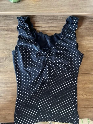 Svart prickig topp  - Snygg svart topp med vita prickar och volangkant vid axlarna. Djupare ringning med liten rosett framtill. Figurnära passform 💕