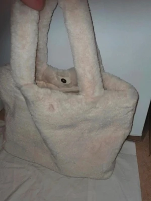 Beige fluffig handväska - Supermjuk handväska i beige fuskpäls med breda handtag och tryckknapp. Perfekt för dig som gillar mysiga accessoarer och vill ha plats för allt du behöver. Väskan har en enkel, fyrkantig form och känns riktigt trendig.