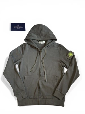 Svart hoodie från Stone Island - Snygg svart hoodie från Stone Island med dragkedja framtill och klassisk logotyp-patch på vänster ärm. Hoodien har huva med dragsko och två fickor framtill. Perfekt för en avslappnad streetwear-look.
