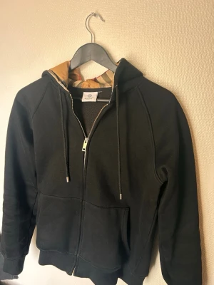 Svart hoodie från burberry  - Svart hoodie med dragkedja och kängurufickor. Huvan är fodrad med ett klassiskt rutigt tyg i beige, rött och svart som är typiskt för burberry. Mjuk bomullskänsla och snören i huvan för justering. Perfekt för en chill och stilren look.