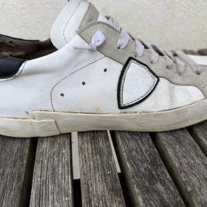 Vita sneakers från Philippe Model - Stilrena vita sneakers från Philippe Model med grå mockadetaljer och svart hälparti. Skorna har en ikonisk sköldlogga på sidan, vita skosnören och perforerade detaljer. Ovandelen är i läder och mocka, sulan är vit och klassisk låg modell. Skorna har sina skavanker men inget synligt när man bär de, ett par fräscha skor som passar till torrt väder! Priset är satt lågt pga skavankerna. Tveka inte att höra av er vid frågor!