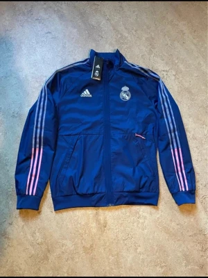 NY Adidas Real Madrid Anthem Jacka - Helt ny och oanvänd Adidas Real Madrid Anthem-jacka i mörkblått med de karaktäristiska rosa och grå ränderna på ärmarna. 