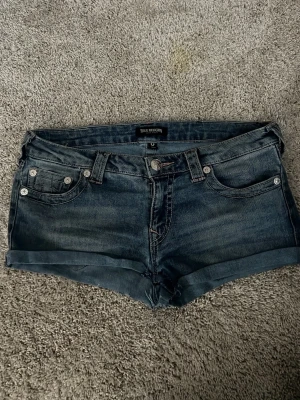 True Religion blå jeansshorts - Blå jeansshorts från True Religion med låg midja, klassiska fem fickor och uppvikta benslut. Snygga kontrastsömmar och ikonisk logga på bakfickan. Tillverkade i mjuk bomull för en bekväm känsla.