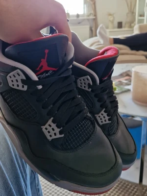 Svarta Air Jordan 4 Retro sneakers - Grymma Air Jordan 4 Retro sneakers i svart läder med röda och grå detaljer. Klassisk Jumpman-logga på plösen och meshpaneler på sidorna. Skorna har högt skaft och cool design som sticker ut. Perfekta för dig som vill ha en ikonisk och streetinspirerad look.