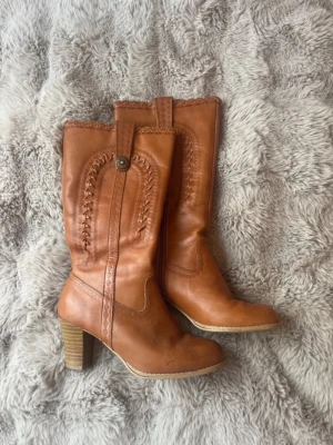 Bruna höga läderstövlar med klack - Snygga bruna stövlar i äkta läder med klack och dekorativa sömmar längs skaftet. Dragkedja på insidan och detalj med knapp upptill. Vätske fläck och andra täcken på användning syns på bild.