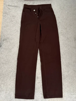 Bruna raka jeans med knäppning - Säljer ett par bruna jeans med raka ben och hög midja. Jeansen har klassisk femficksdesign och stängs med synliga silverfärgade knappar framtill. Materialet är kraftig denim i bomull. Perfekta för en stilren och avslappnad look.
