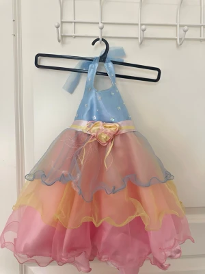 Pastellfärgad klänning  - Superfin prinsessklänning i pastellfärger med blå överdel, dekorativa pärlor och volangkjol i rosa, gul och blå. Klänningen har halterneck och rosett i midjan. Den är storlek 0-18 månader💗💗