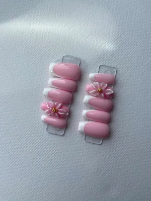 Rosa press-on naglar med blommor - Snygga press-on naglar i ljusrosa med vit fransk tipp och 3D-blommor med gulddetalj. Naglarna har en oval form och ger en söt och trendig look. Perfekt för dig som vill ha en feminin och unik nageldesign.