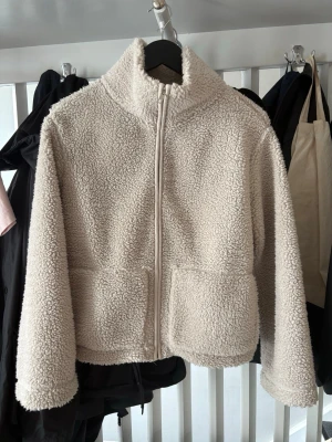Beige teddyjacka med dragkedja - Mysig beige teddyjacka med hög krage och hel dragkedja framtill. Jackan har två stora fickor framtill och är tillverkad i fluffigt teddy-material som ger en avslappnad och trendig look. Perfekt för kyliga dagar när du vill vara både varm och snygg.