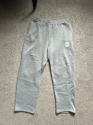 Grå mjukisbyxor med tryck - Säljer ett par ljusgrå mjukisbyxor med resår i midjan och ett vitt tryck med bokstäver på vänster lår. Byxorna har raka ben och är gjorda i mjukt, bekvämt material som passar perfekt för chill dagar.